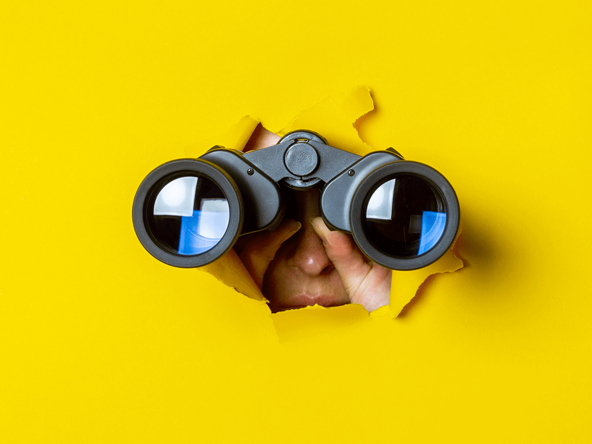 Binoculars are see bursting through a yellow paper background. We can see that a person is holding these and looking through the viewfinder. The image is used to depict searching and is used alongside job searching .